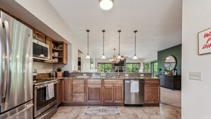 Una cocina moderna con encimeras de granito, electrodomésticos de acero inoxidable, armarios de madera e iluminación colgante, con vistas al exterior.