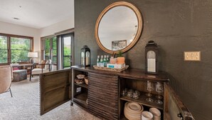 Una habitación con una pared oscura, una consola de madera con un espejo, estantes con copas y una silla.