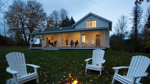Una casa con porche, tres sillas Adirondack blancas y un brasero.
