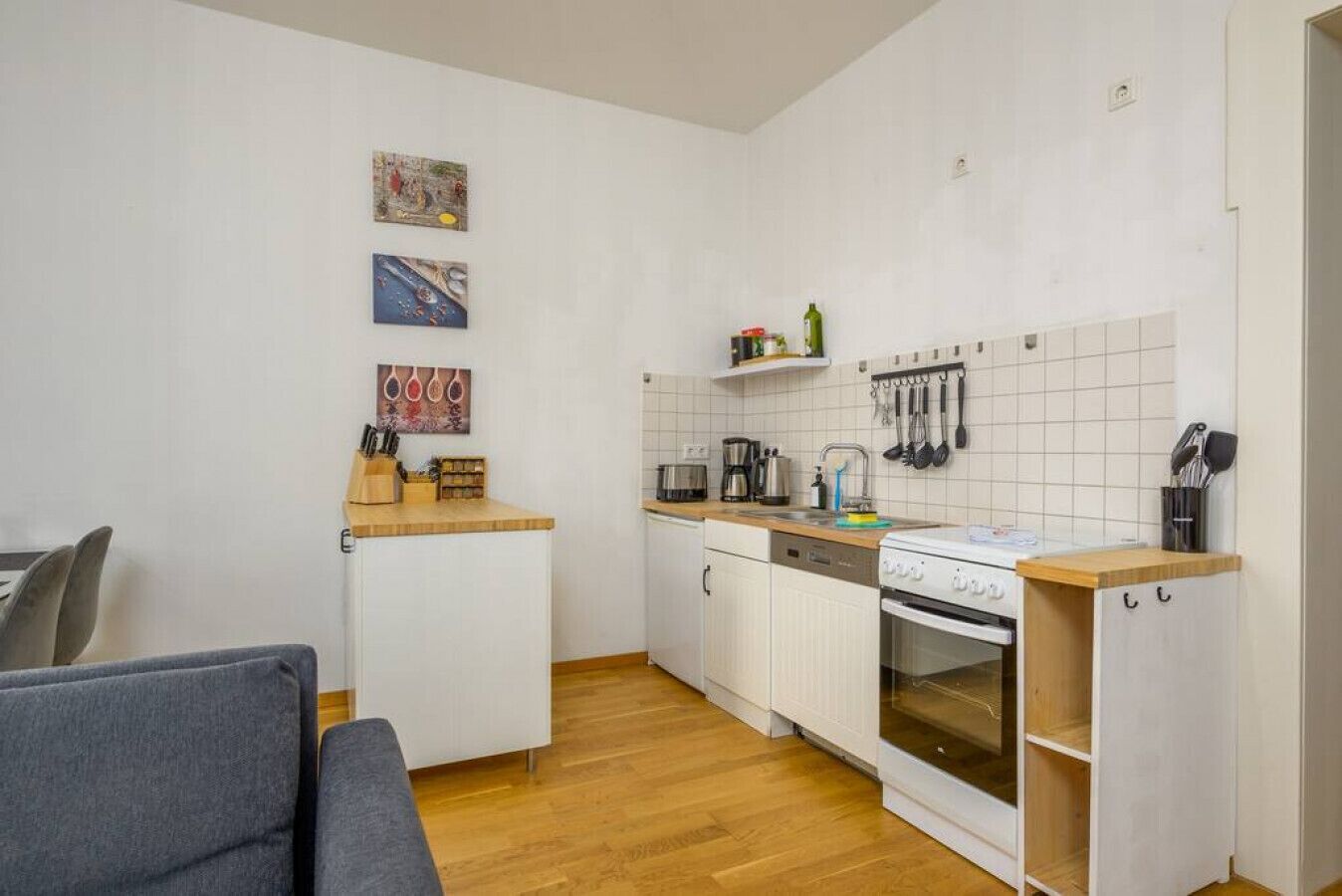 Een kleine keuken met witte kastjes, een houten aanrechtblad en een fornuis. Boven de gootsteen bevinden zich twee wandplanken met keukengerei.