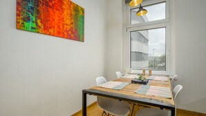 A dining area with a wooden table, four chairs, a colorful abstract painting, and a large window with a view of buildings.