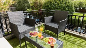 Un balcon agrémenté de meubles en osier, une table en verre avec des fruits et des boissons, et une vue sur un jardin et un bâtiment en arrière-plan.