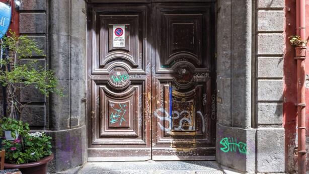 Un arc en pierre mène à une porte en bois sombre, recouverte de graffitis, et sur laquelle est apposé un panneau indiquant « Stationnement interdit ».
