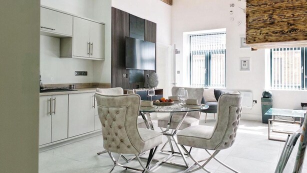 A modern kitchen with white cabinets, a large TV, and a dining area with a glass table and cushioned chairs. - Blissful Bradford Apartment - City Center Gem (Bradford)