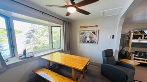 Ein Zimmer mit Holztisch und Bank, Deckenventilator, Fenster mit Blick auf Bäume, einem Bärenbild an der Wand und Etagenbetten in der Ecke.