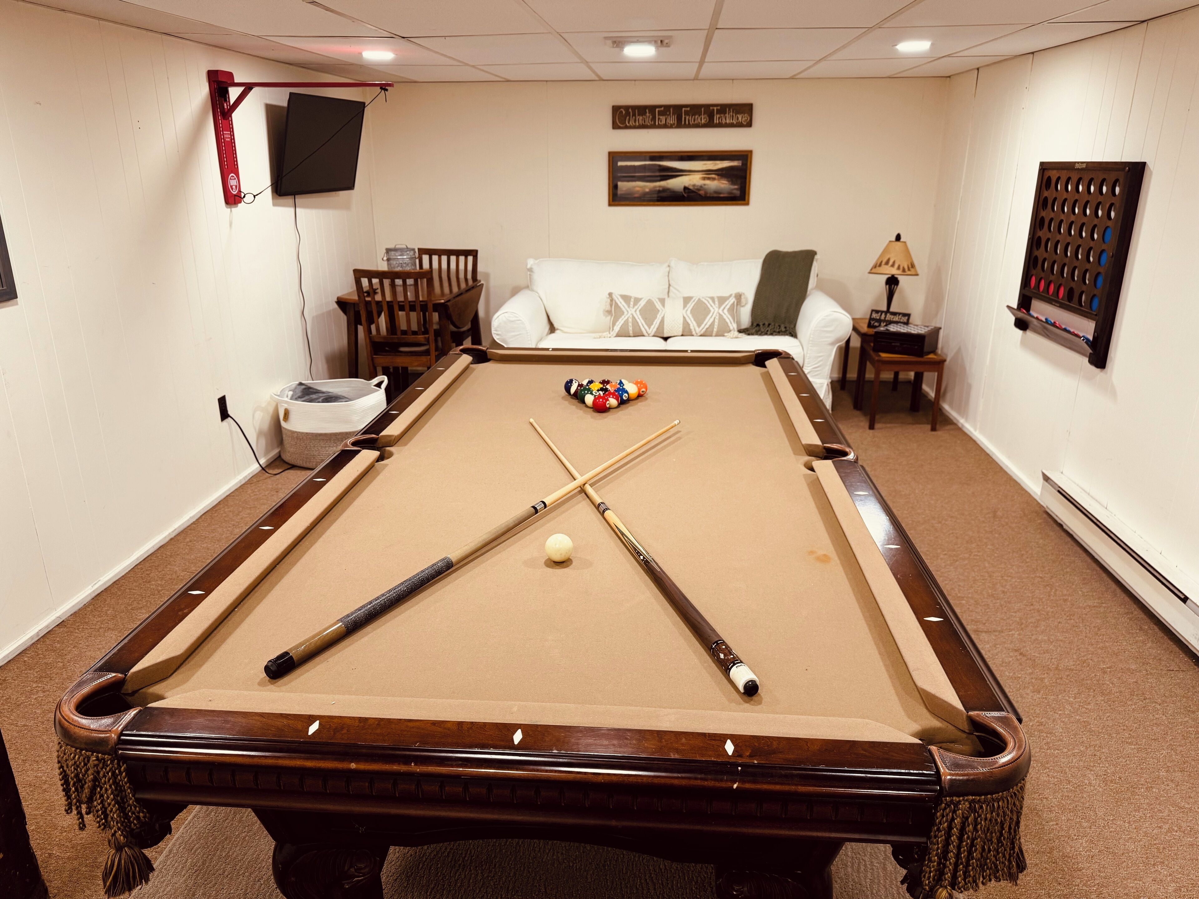 A pool table with two cues and a billiard ball, a white sofa, a small table with a lamp, and a framed picture on the wall.