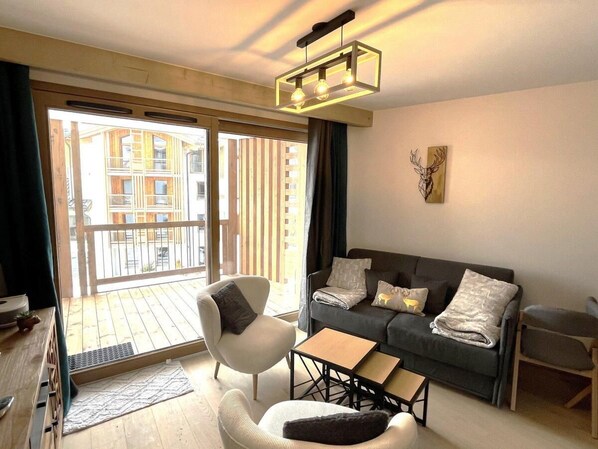 A modern living room with a sofa, armchair, and coffee table, a balcony with wooden railings, and a cityscape view.
