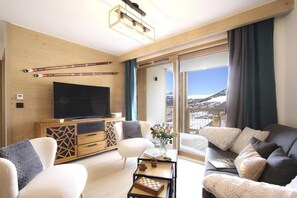 A modern living room with a large flat-screen TV, wooden furniture, and a view of a snowy landscape through a sliding glass door.