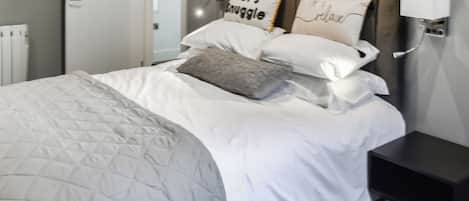 A neatly made bed with a quilted grey duvet, two pillows with text, and a bedside table with a lamp.