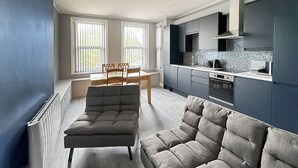 A modern living room with a sectional sofa, a dining table with chairs, and a kitchen area with dark cabinets and a tiled backsplash. - Minutes from Folkestone Central Station and modern flat with three bedrooms. (Cheriton, near Folkestone)