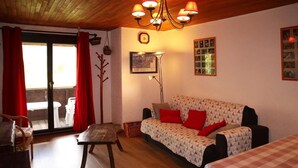 Ein gemütliches Wohnzimmer mit Holzbalkendecke, einer Couch mit gemusterten Kissen, einem Esstisch mit kariertem Tischläufer und Blick durch eine Glastür.