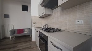 A compact kitchen with a gas stove, oven, and white cabinets. There is a small window above the sink and a radiator in the corner.