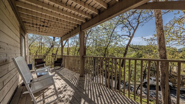 Una terraza de madera con un pérgola, asientos exteriores y vistas a árboles y una carretera.