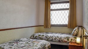 Et lille, hyggeligt værelse med to senge, et vindue med gardiner, et natbord med lampe og en radiator.
