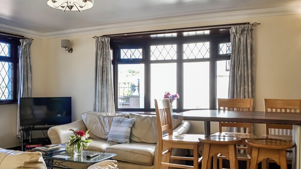 En stue med sofa, sofabord, fjernsyn, spisebord med stole og store vinduer med gardiner.