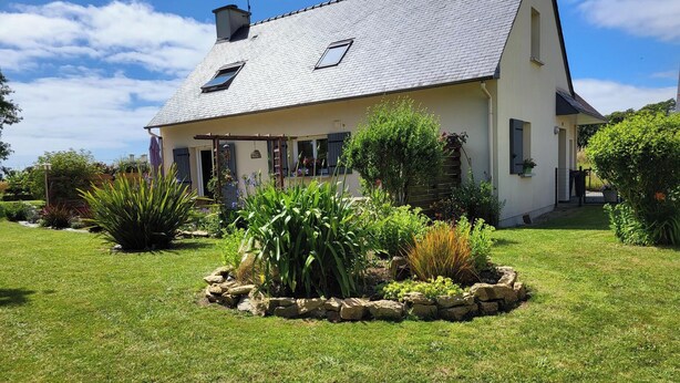 A two-story house with a sloped roof, a small porch, and a garden with various plants and a stone border. - Private room, in a quiet, leafy setting.  (ploneour lanvern)