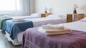 Ein Hotelzimmer mit drei Betten, jedes mit einer andersfarbigen Decke. Zwei Nachttische mit Lampen sind vorhanden.