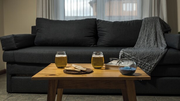 Ein dunkelgraues Sofa mit Decke, zwei Biergläser, eine Schale mit Snacks und ein Buch auf einem Holztisch davor.
