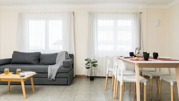 Una sala moderna con un sofá gris, una mesa de centro de madera y una zona de comedor con una mesa y sillas blancas.