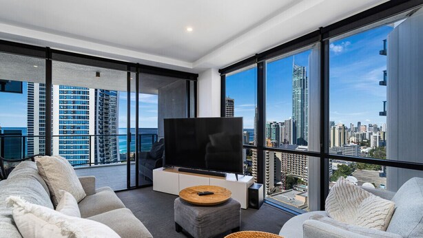 Una sala de estar moderna con un televisor de pantalla plana grande, asientos cómodos y vistas a un horizonte urbano a través de ventanales de piso a techo.