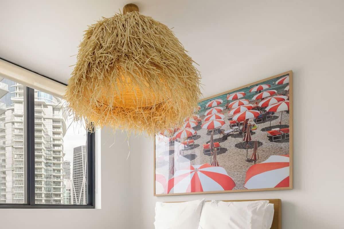 A bedroom with a bed, a hanging light fixture, a painting of beach umbrellas, and a view of tall buildings through a window.