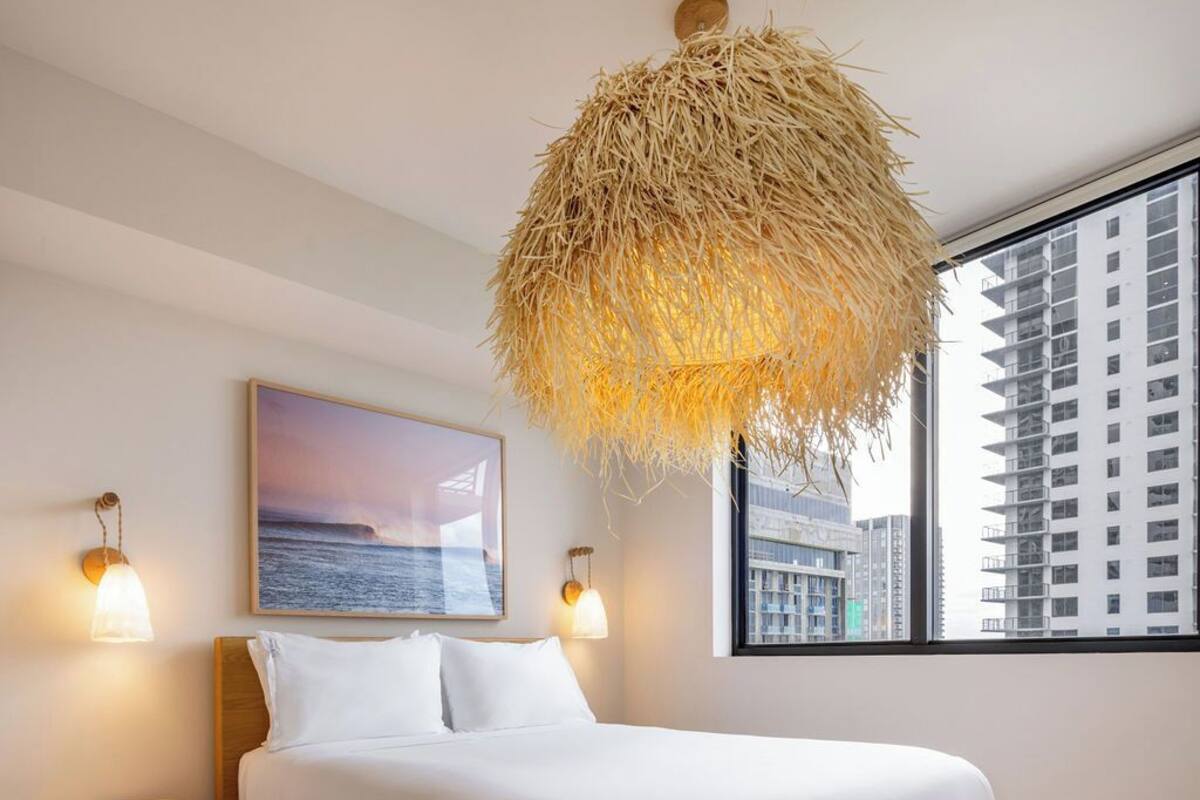 A bedroom with a large bed, a hanging light fixture, a painting, and a view of buildings through a window.
