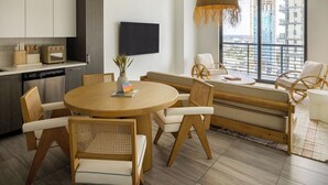 A modern living room with a round wooden dining table, white cushioned chairs, a woven hanging light fixture, and a large window offering a city view. - Flow Hotel Miami (Miami)