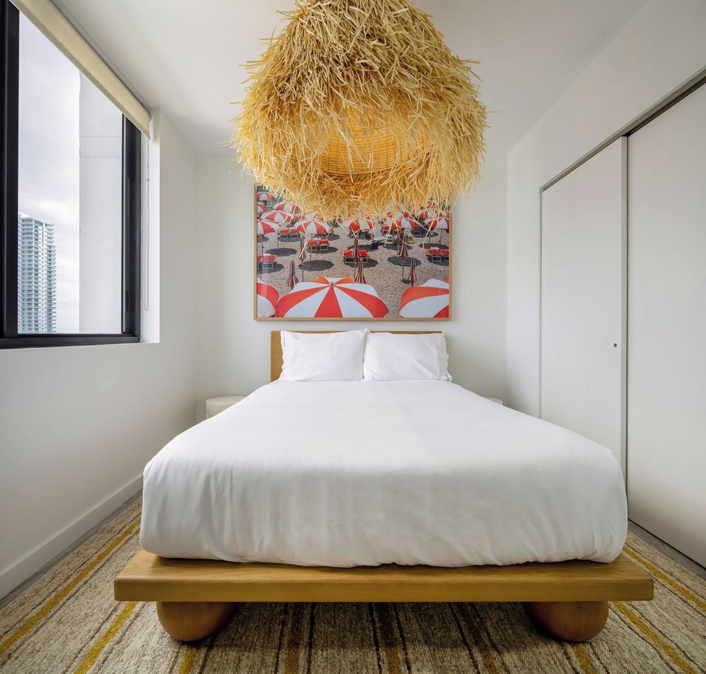A modern hotel room with a large bed, a straw chandelier, a beach-themed painting, and a view of the city through a window.