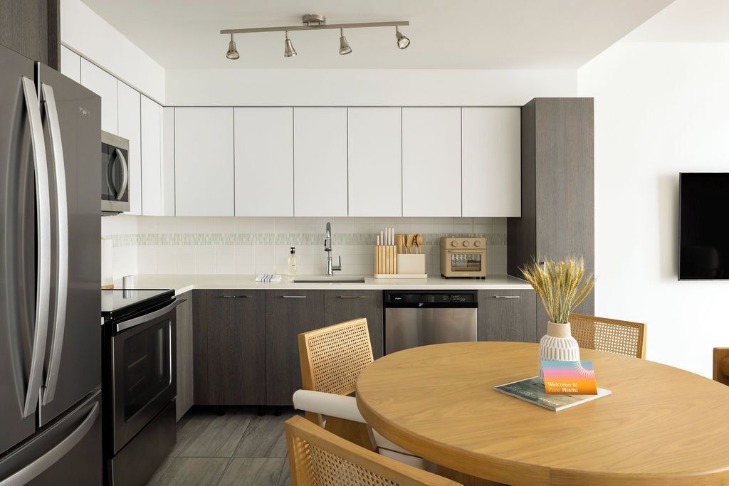 Cocina moderna con electrodomésticos de acero inoxidable, armarios blancos y una mesa de comedor de madera con un letrero de bienvenida.