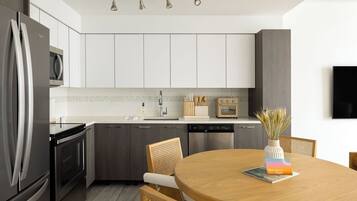 Cocina moderna con electrodomésticos de acero inoxidable, armarios blancos y una mesa de comedor de madera con un letrero de bienvenida.