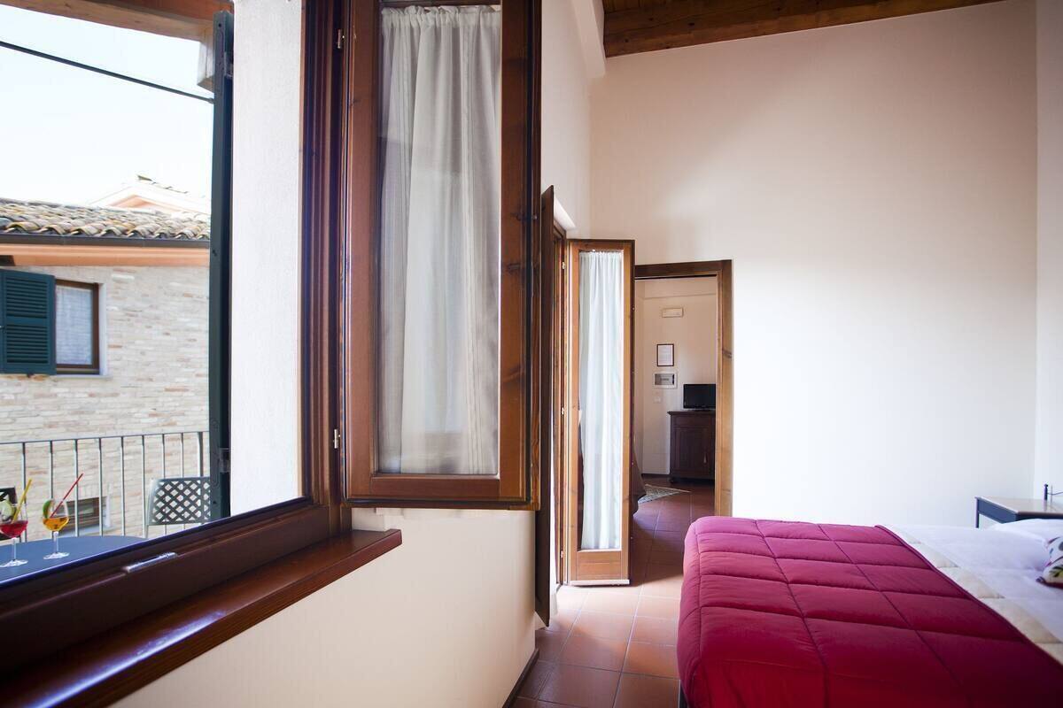 A bedroom with a red bed, a window with a curtain, a balcony, and a view of a building outside.