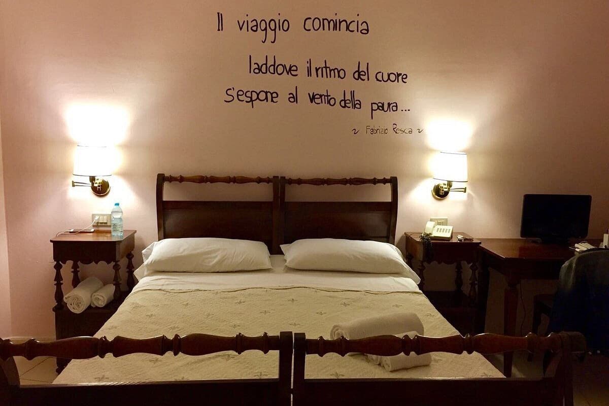 A neatly made bed with a headboard, two bedside tables with lamps, a desk with a computer, and a wall with a quote in Italian.