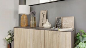 Ein Holzschrank mit Spiegel darüber, der ein gerahmtes Schild mit dem Aufdruck 'CITY', eine weiße Vase und eine Korbtaschelampe zeigt.