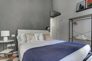 A modern hotel room with a large bed, a bedside table with a lamp, a small side table, and a wardrobe with a hanging rack. - Stylish Apartment in Historic Rione Celio (Roma)