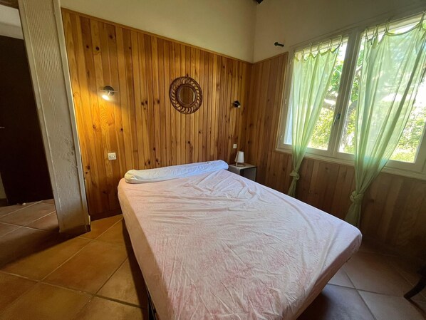 Une chambre avec un lit, des murs recouverts de panneaux de bois, un miroir fixé au mur, une fenêtre avec des rideaux et une petite table de chevet.