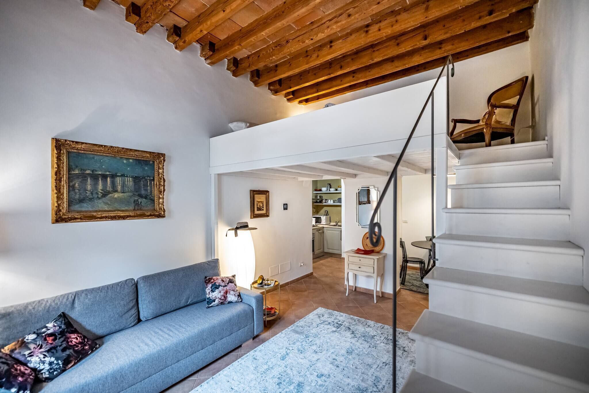 A living room with a sofa, a painting, a staircase, and a kitchen in the background.