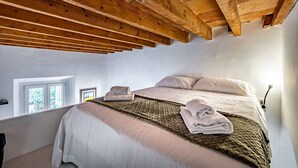 A bedroom with a high ceiling, a bed with a quilt, towels, a small window, and a wall-mounted lamp.