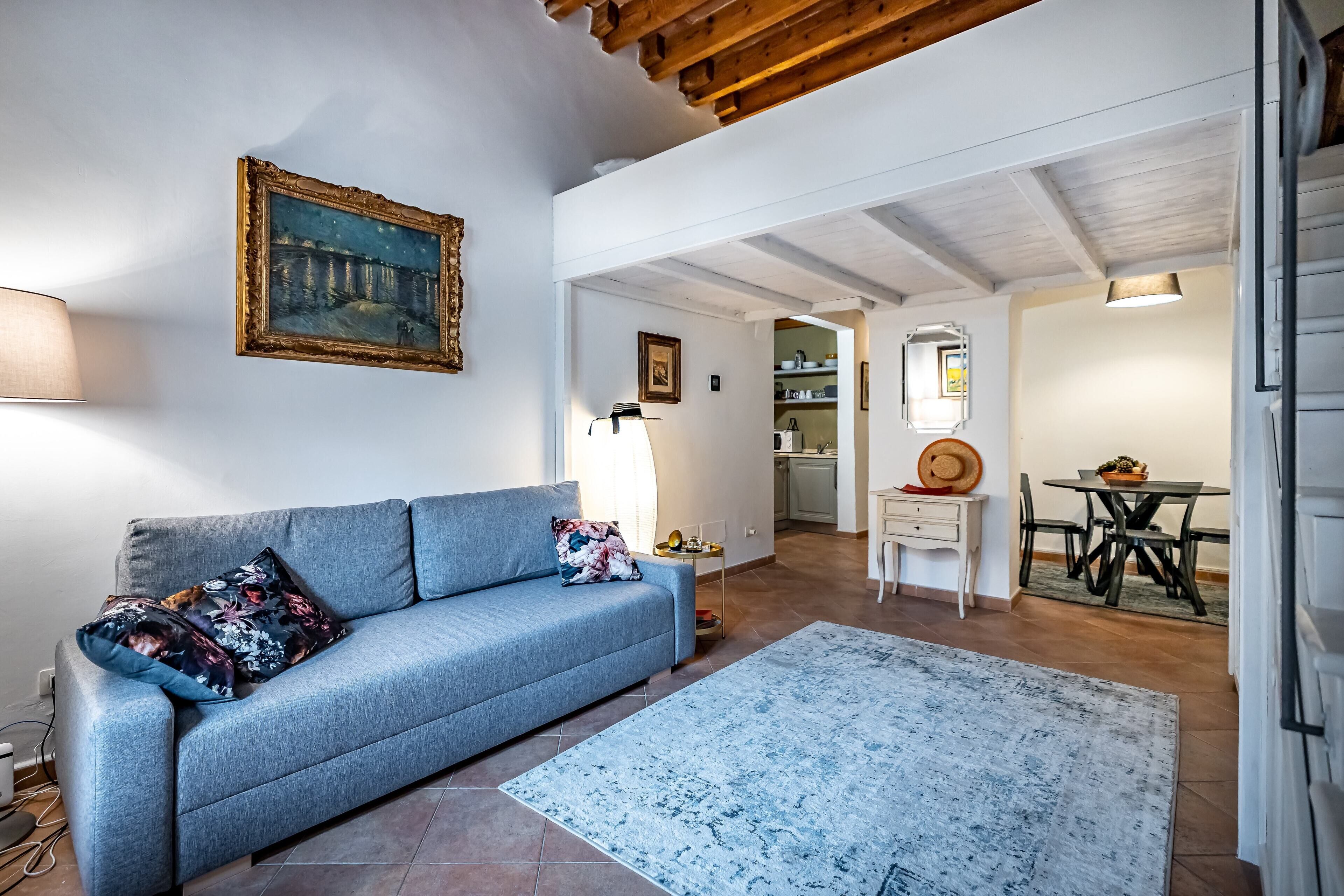 A living room with a sofa, a rug, a painting, and a dining area with a table and chairs.