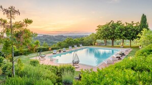 Une piscine rectangulaire, agrémentée de chaises longues et d’un fauteuil suspendu, entourée d’une végétation luxuriante et d’arbres, et offrant une vue sur des collines lointaines.