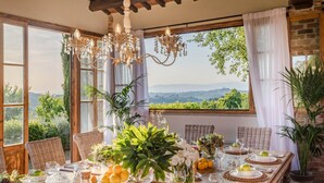 Une table à manger dressée avec des assiettes, des verres et des fruits, entourée de chaises en osier, et offrant une vue sur la verdure et les montagnes à travers de grandes fenêtres.