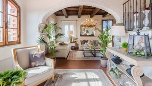 Un vaste salon doté d’un plafond haut, de poutres en bois et d’une porte en arc de cercle. On y trouve plusieurs coins salon, un lustre et divers objets décoratifs.
