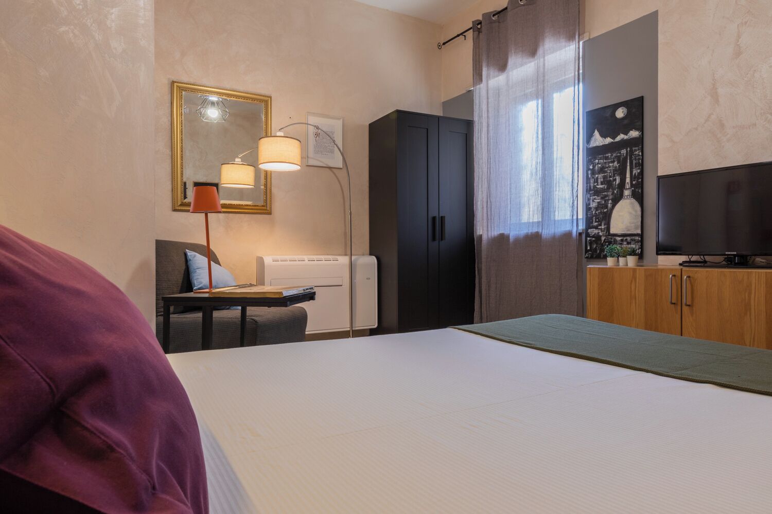 A hotel room with a bed, a desk with a lamp, a wardrobe, a radiator, and a television.