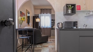 Eine kompakte Küche mit Mikrowelle, Wasserkocher und Kaffeemaschine. Ein Essbereich mit Tisch und Stühlen sowie ein Sofa sind vorhanden.