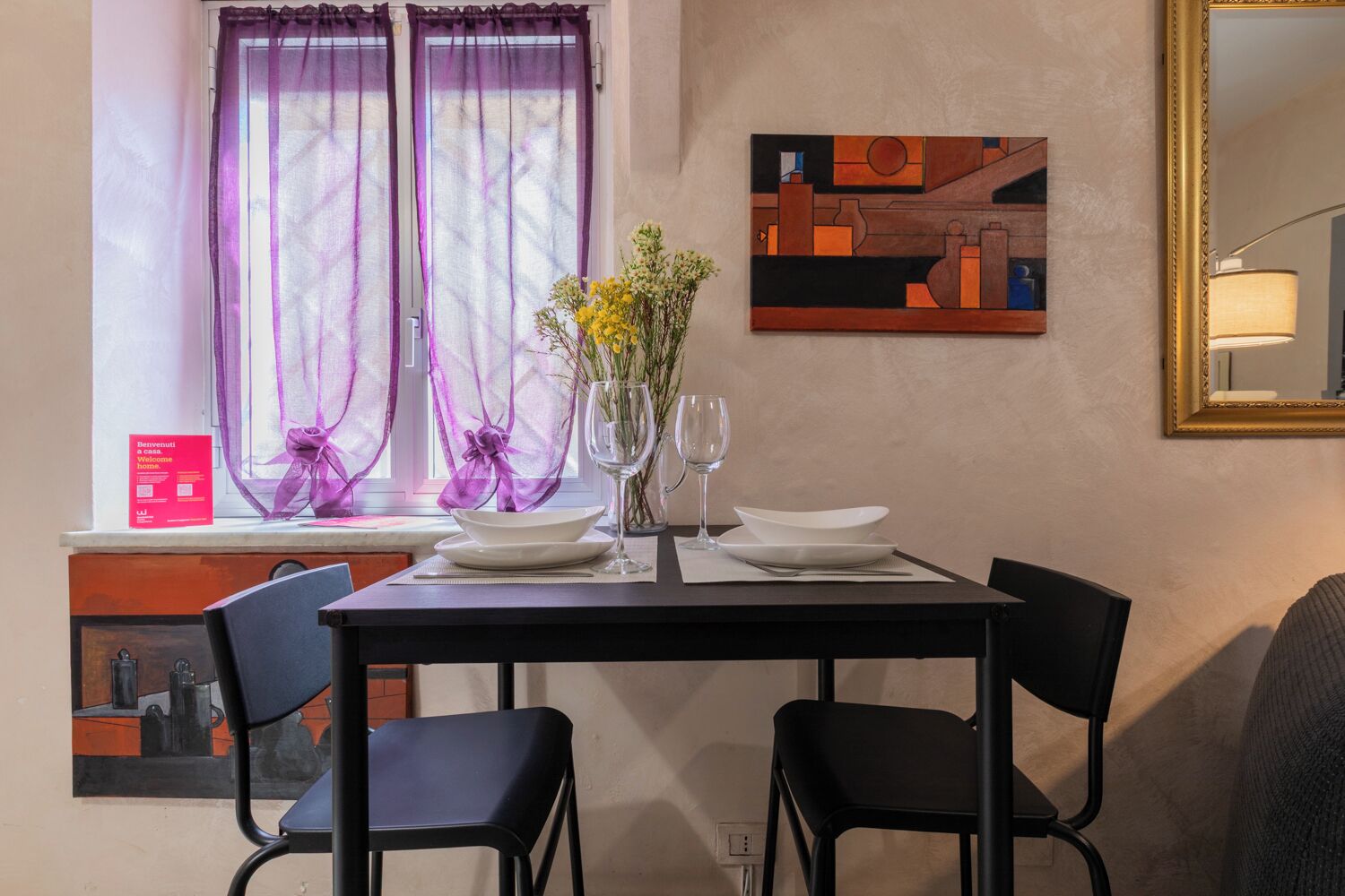Un coin repas avec une table dressée pour deux personnes, un vase rempli de fleurs et un tableau accroché au mur.