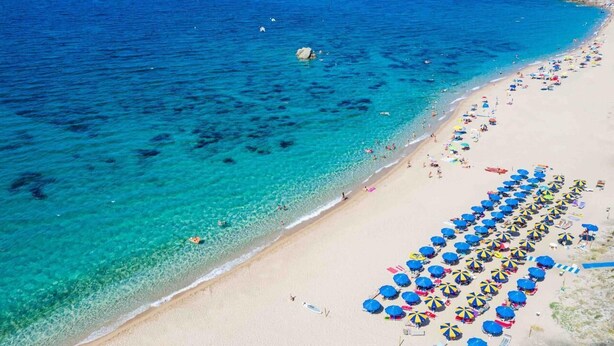 Vista aérea de una playa con aguas turquesas cristalinas, una orilla arenosa y numerosos sombrillas coloridas.
