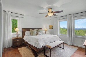 Un dormitorio con cama, mesitas de noche, un banco, ventilador de techo y ventanas amplias con vistas.