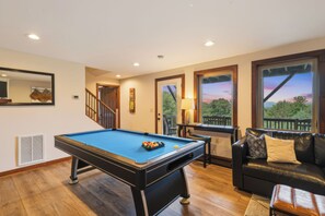 Une table de billard dans une pièce dotée d’un plancher en bois, d’un canapé en cuir et de grandes fenêtres offrant une vue dégagée.