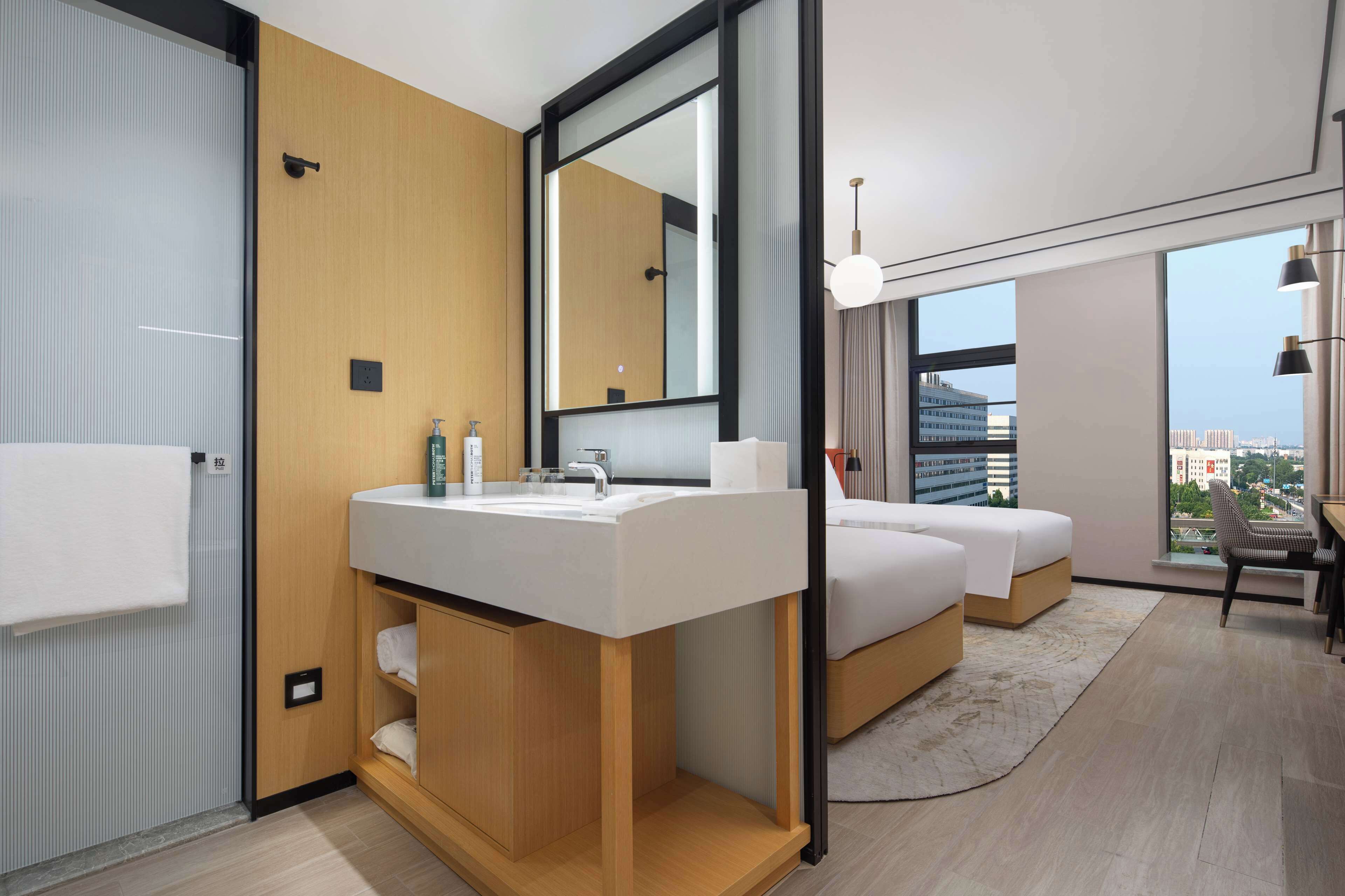 Ein modernes Hotelzimmer mit großem Fenster, einem Holzanrichte mit Waschbecken, zwei Betten und einem Schreibtisch mit Stuhl.