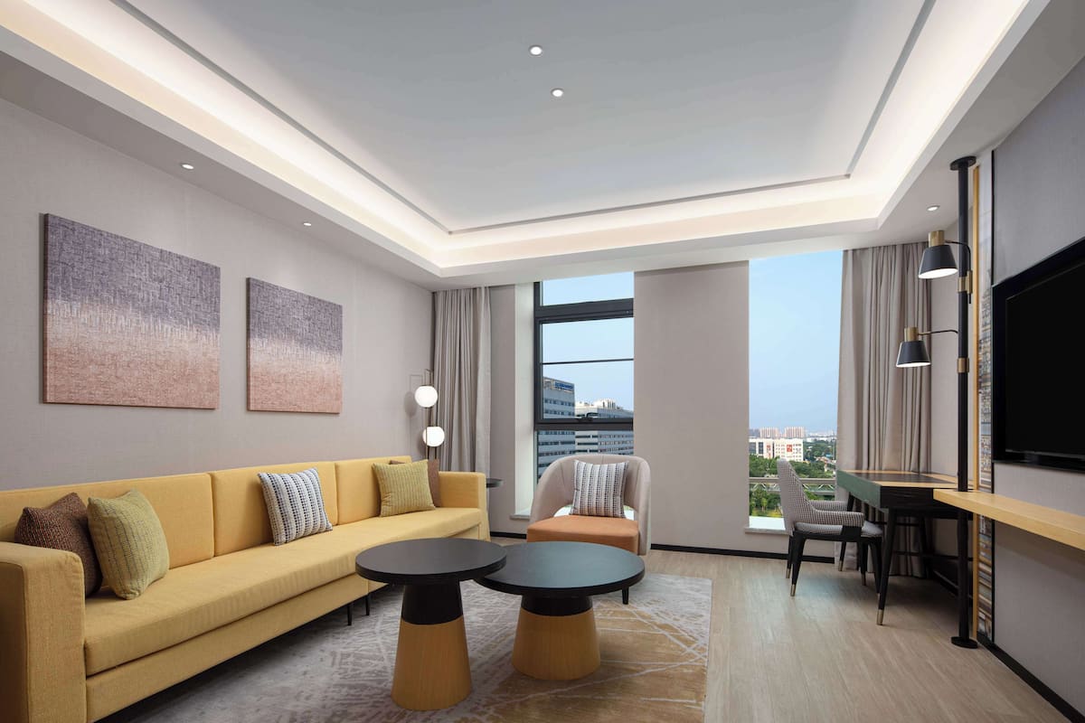 Une chambre d’hôtel moderne avec un canapé, deux petites tables rondes, un téléviseur et une vue sur la ville grâce à de grandes fenêtres.