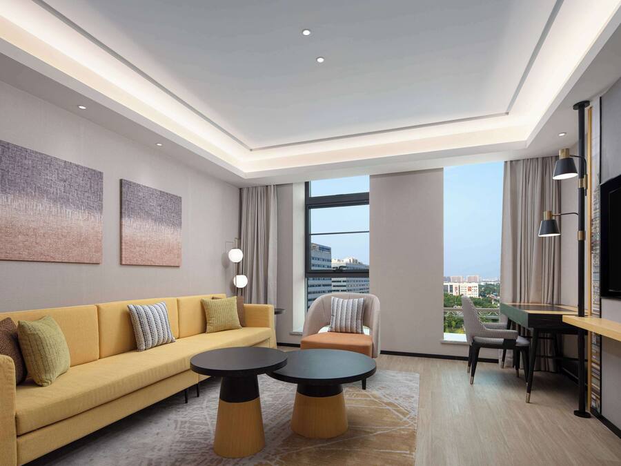 A modern hotel room with a sofa, two round tables, a TV, and a city view through large windows.